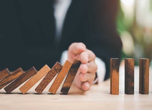 Hand stopping a row of falling wooden domino blocks on a table, symbolizing risk management and prevention.