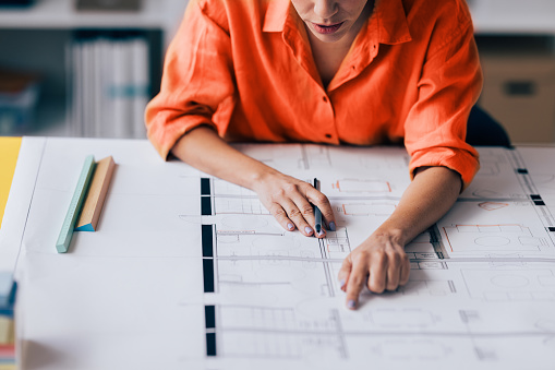 Person reviewing and pointing at architectural blueprints on a desk, with drafting tools nearby.
